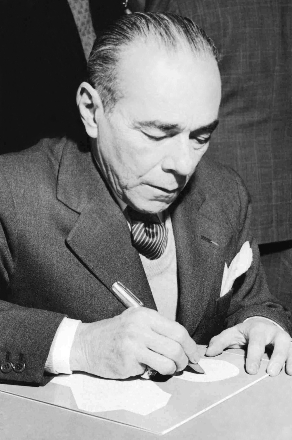 Fotografia em preto e branco. Homem de cabelo lisos para trás, terno escuro. Ele segura uma caneta e escreve em uma folha apoiada sobre uma superfície.