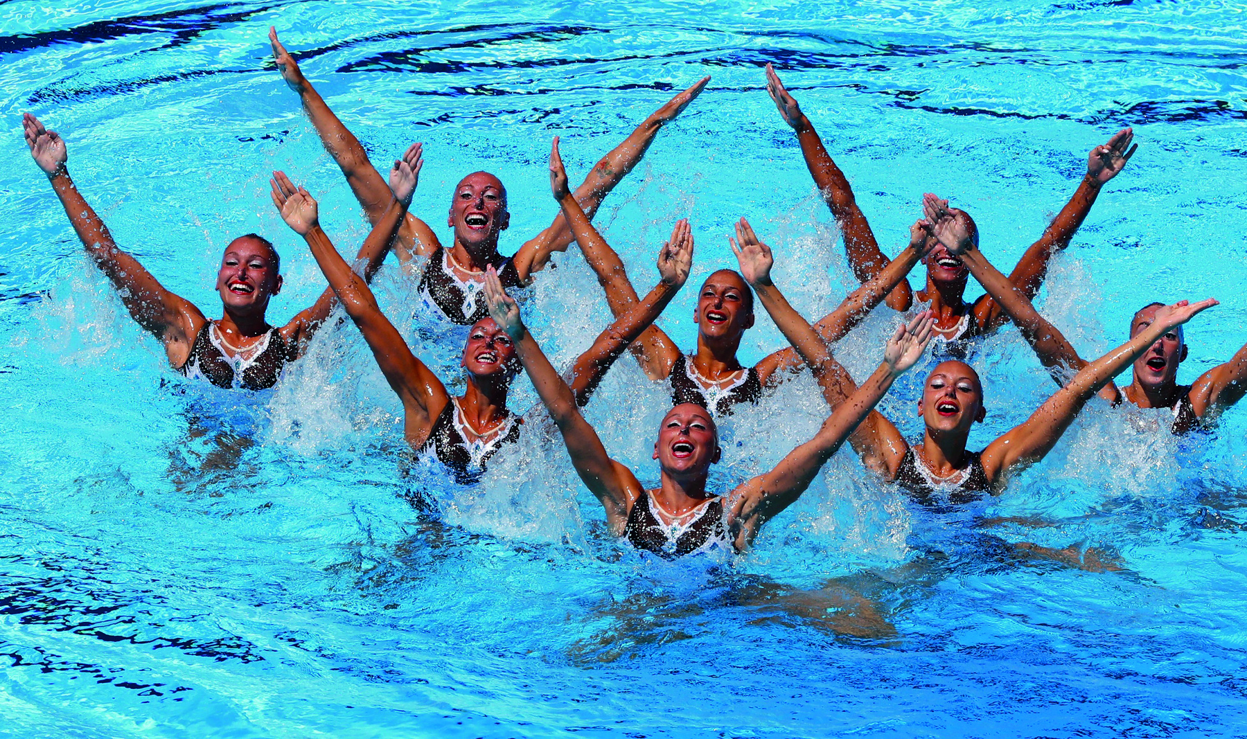 Fotografia. Oito mulheres em uma piscina, todas com o tronco do corpo para fora da água e seus braços abertos para cima.