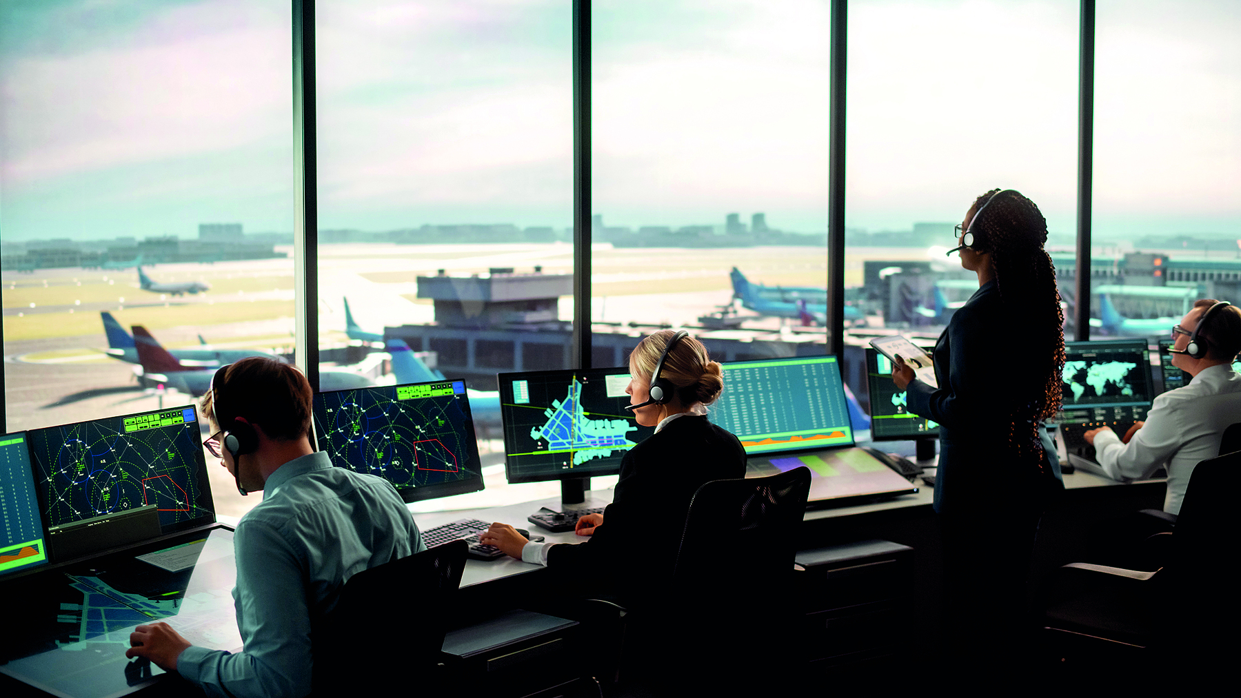 Fotografia. Vista parcial de um torre de controle aéreo. Algumas pessoas estão sentadas à frente de monitores com informações na tela. Ao fundo, pista de pouso e aviões.