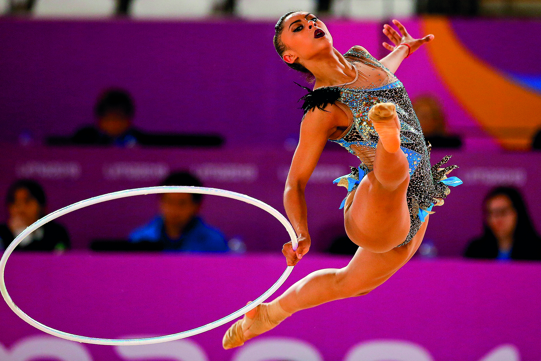 Fotografia. Vista frontal de ginasta de collant no ar com as pernas abertas e os braços para o lado. Ela segura um aro branco, como um bambolê, próximo às pernas.