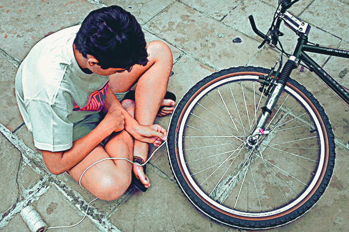 fotografia. Vista de cima de garoto sentado no chão. Ao lado, parte de uma bicicleta com destaque para o pneu frontal dela. Ele segura um barbante que está ao redor do pneu.