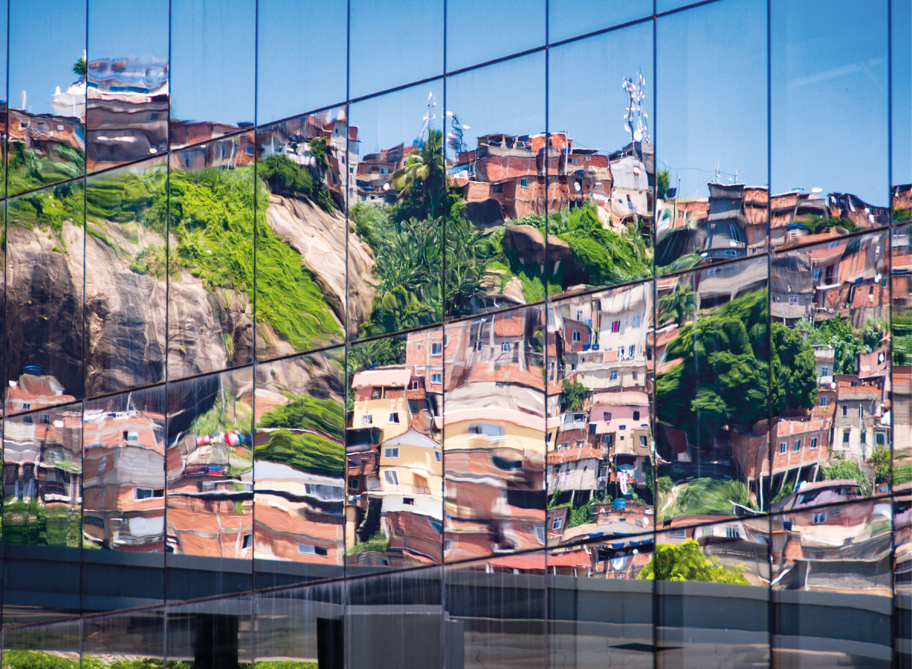 Fotografia. Vista frontal de prédio composto por vidros espelhados refletindo a imagem de um morro com casas de tijolos e abaixo, uma ponte. No centro, as imagens nos espelhos estão um pouco distorcidas.