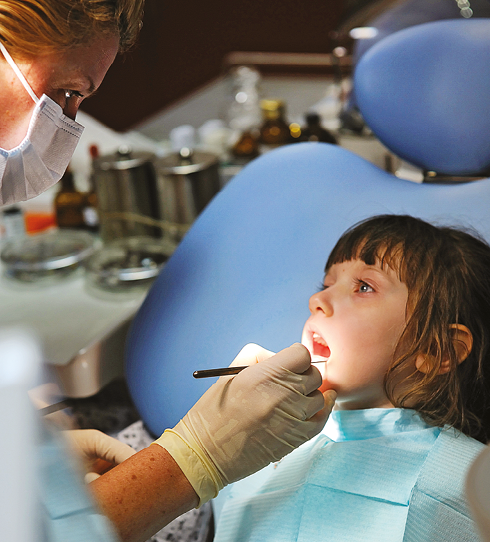 Fotografia. Menina sentada com a boca aberta na cadeira de um dentista. Uma mulher com máscara descartável sobre a boca está com um instrumento examinando a boca da menina.