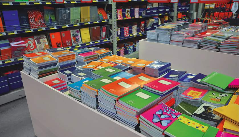Fotografia. Vista superior de uma bancada com diversos cadernos coloridos. Ao fundo, prateleiras com objetos.