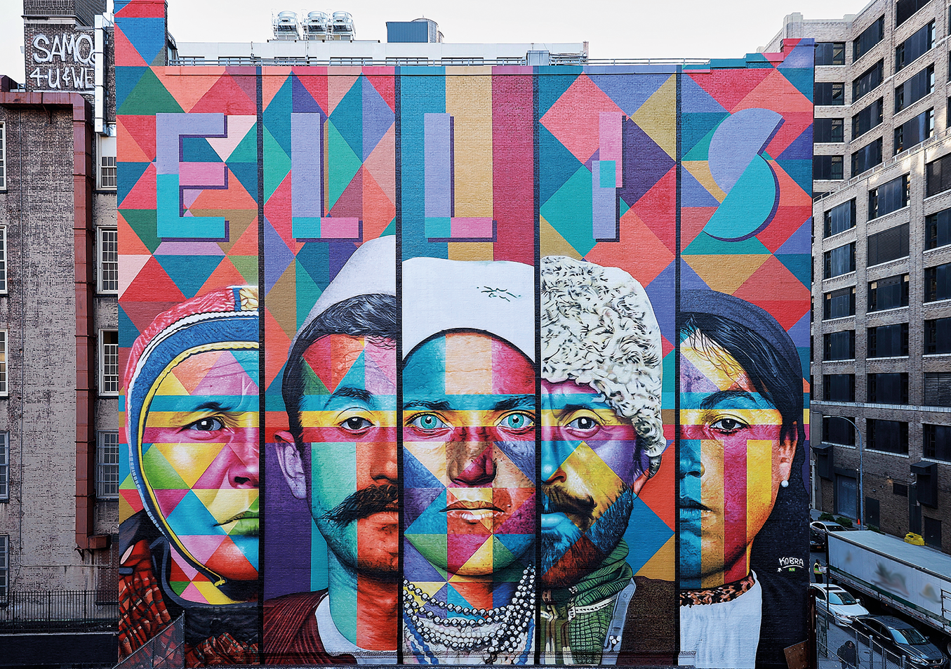 Fotografia. Painel colorido na lateral de edifício com o nome ELLIS acima e abaixo, rosto de 5 pessoas entre homens e mulheres. À direita, rua com carros e construções.