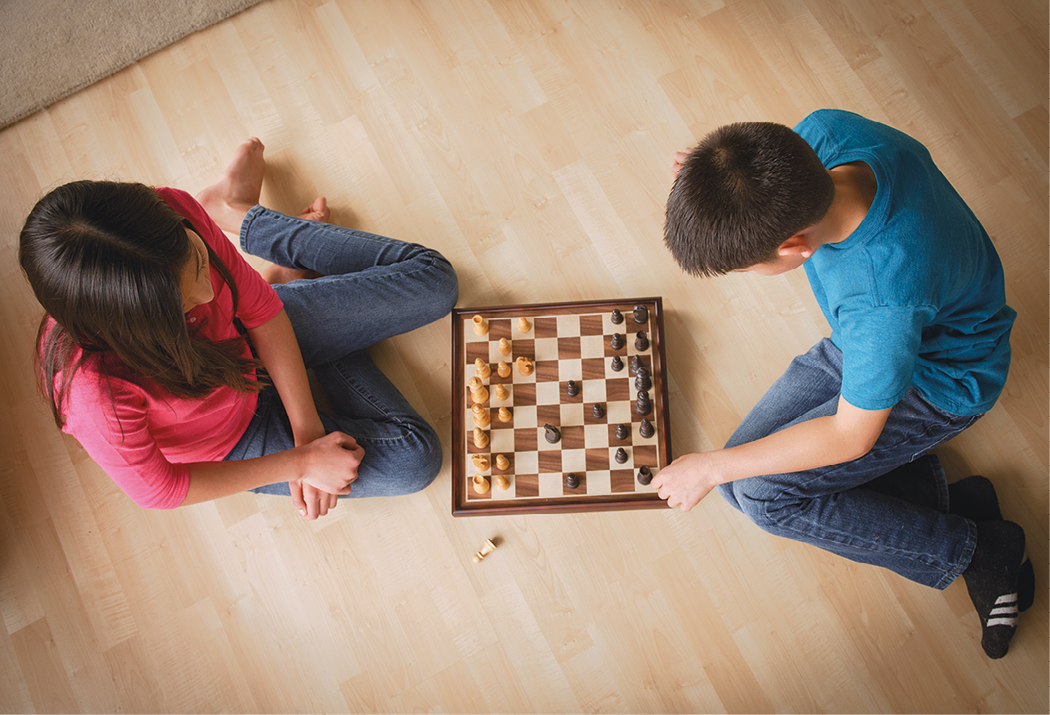Fotografia. Vista do alto de um menino e uma menina sentados no chão jogando xadrez em tabuleiro quadriculado.