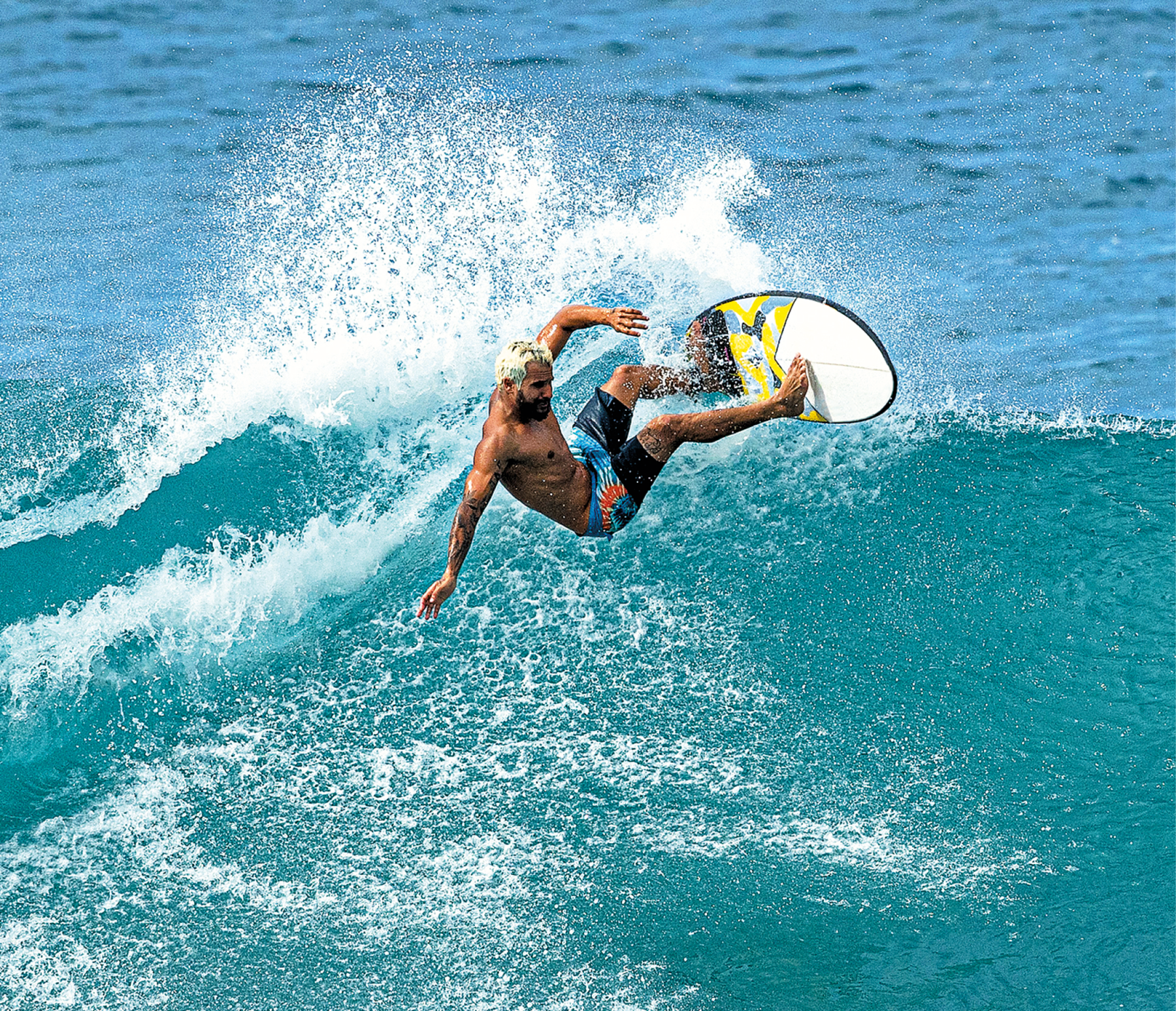 Fotografia. Vista do alto de um homem de bermuda sobre uma prancha de surfe, fazendo uma manobra sobre uma onda no mar.