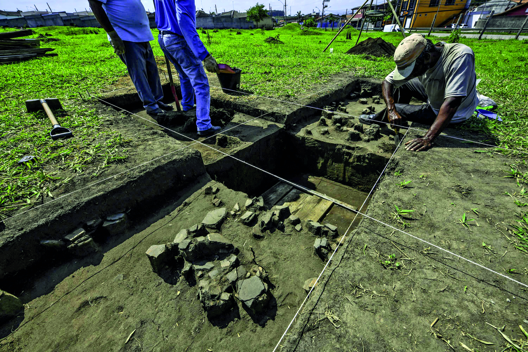 Fotografia. Uma área de escavação arqueológica. Percebe-se que os blocos de escavação são feitos em medidas retangulares. Dois homens estão em pé em um dos retângulos à esquerda e um terceiro homem está abaixado escavando em um retângulo mais à direita. A vegetação ao redor é rasteira.