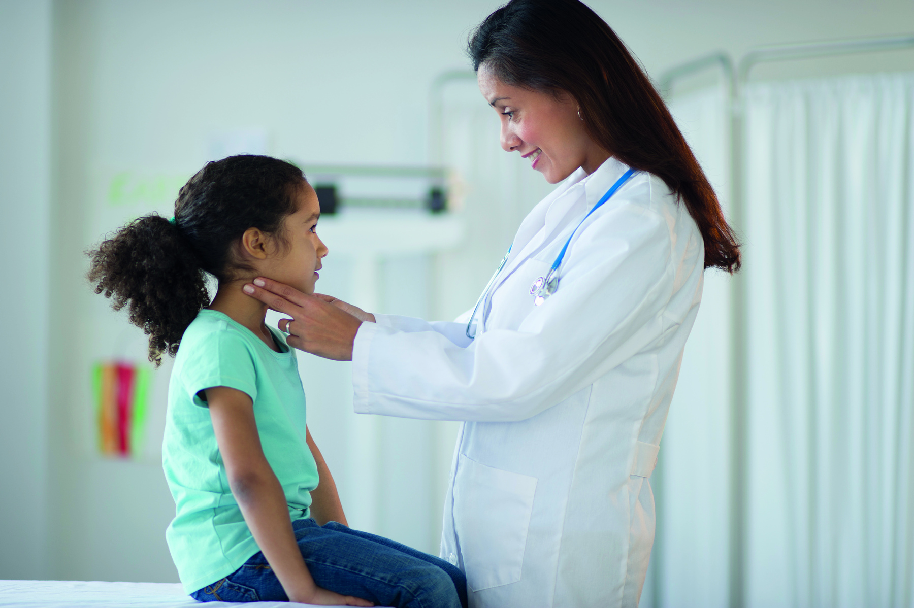 Fotografia. Médica pediatra de cabelo preto, jaleco branco e estetoscópio no pescoço está com as mãos no pescoço de uma menina de cabelo preto e camiseta azul sentada em uma maca.