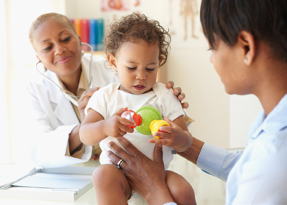 Fotografia. Uma médica negra examina uma criança, ela está com o estetoscópio nas costas da criança, que está segurando um brinquedo. A mãe, que é negra, apoia a criança.