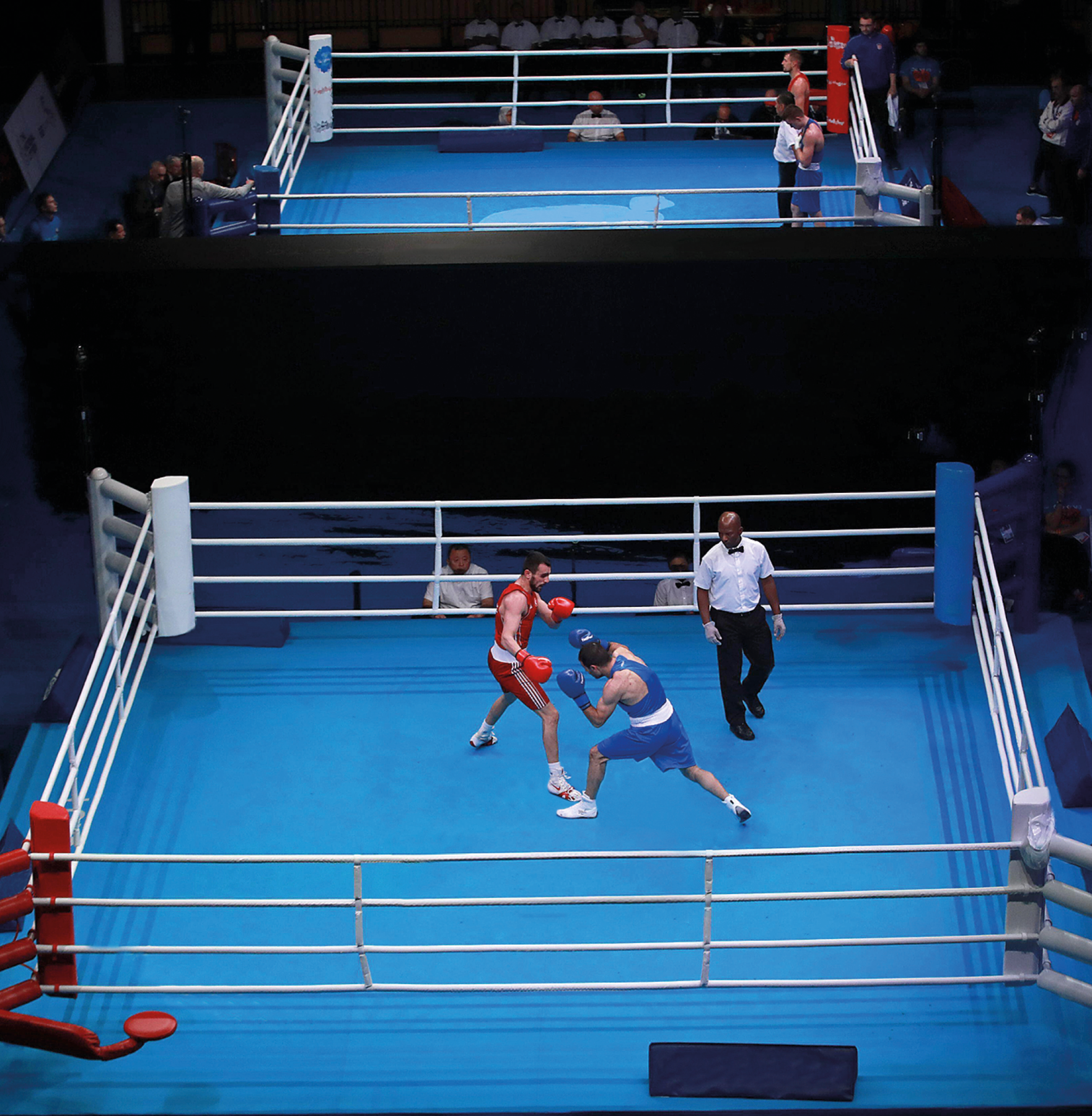 Fotografia. Vista de alto de um ringue de boxe com dois lutadores e um juiz. Ao lado do ringue, duas pessoas assistem a luta. No ringue, à esquerda, homem branco, usando regata vermelha e shorts vermelho. À direita, homem branco, usando regata azul e shorts azul. Ambos usam luvas de boxe. Ao lado deles, homem negro, usando camisa branca, calça preta e gravata borboleta observa os lutadores.