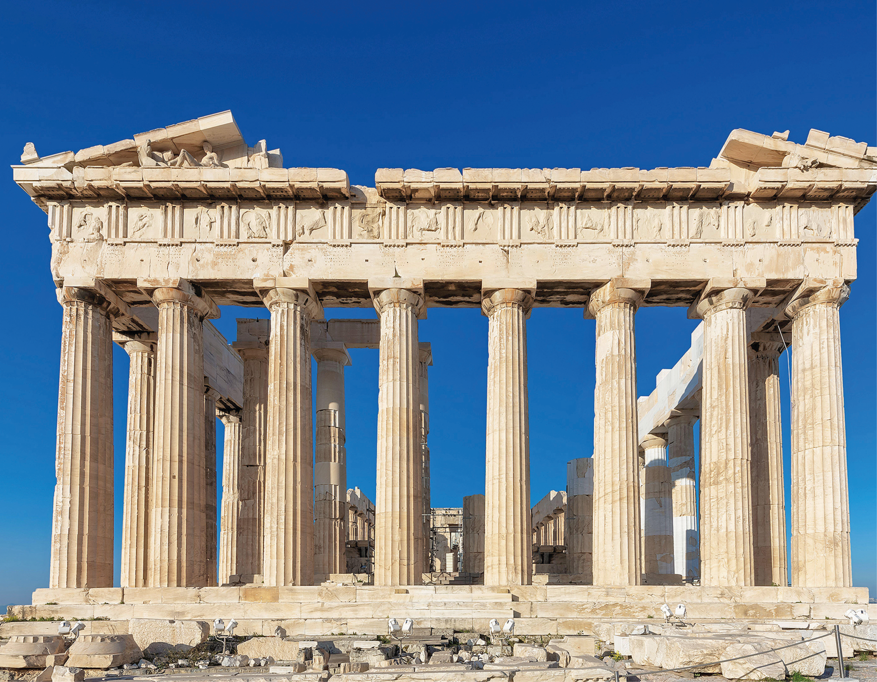 Fotografia. Vista frontal do Partenon, construção grega em ruínas com colunas cilíndricas e degraus. Céu azul sem nuvens.