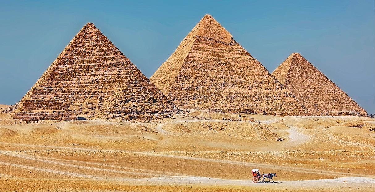Fotografia. Vista frontal de três pirâmides de cor marrom em chão de areia.