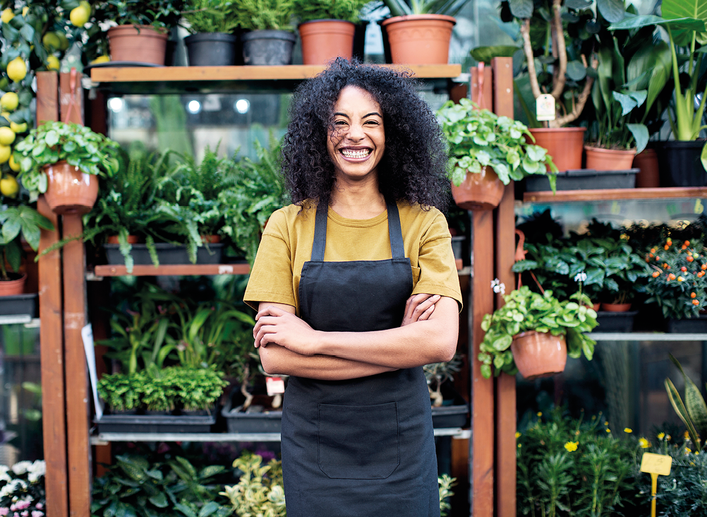 Fotografia. Mulher negra, de cabelos pretos encaracolados, camiseta amarela e avental preto. Ela está sorrindo, em pé com os braços cruzados. Atrás dela, prateleiras com vasos com plantas.