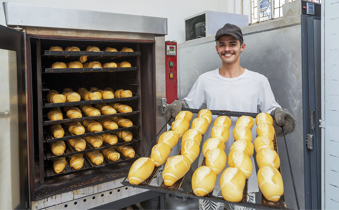 Fotografia. Homem branco, de chapéu, camiseta branca e luvas. Ele segura uma bandeja com pães. Ao lado, forno com pães.