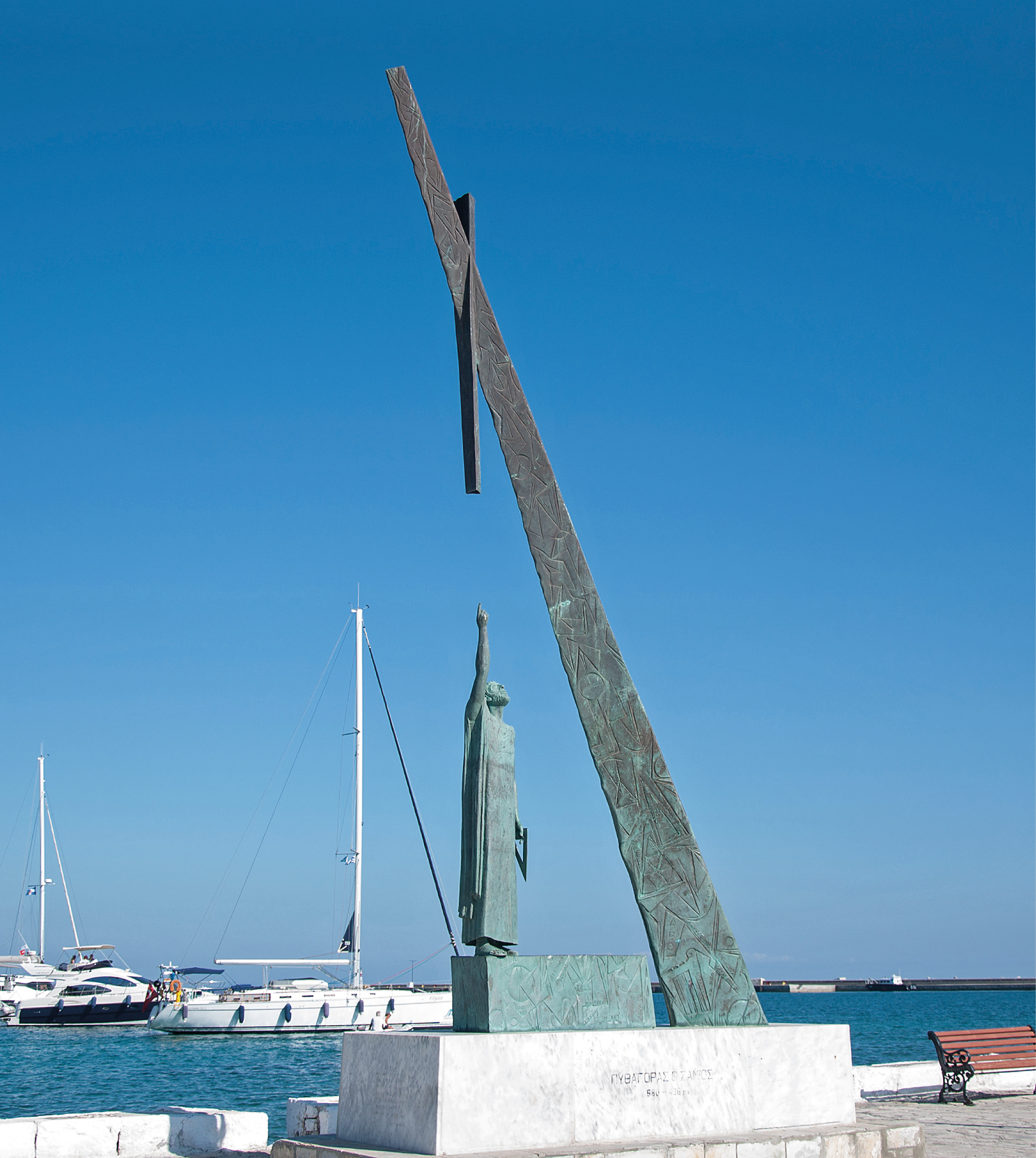 Fotografia. Base com formato de bloco retangular e sobre ela um monumento de bronze de uma pessoa em pé com um dos braços para cima onde há uma haste presa a outra haste na diagonal. Ao fundo, barcos no mar.