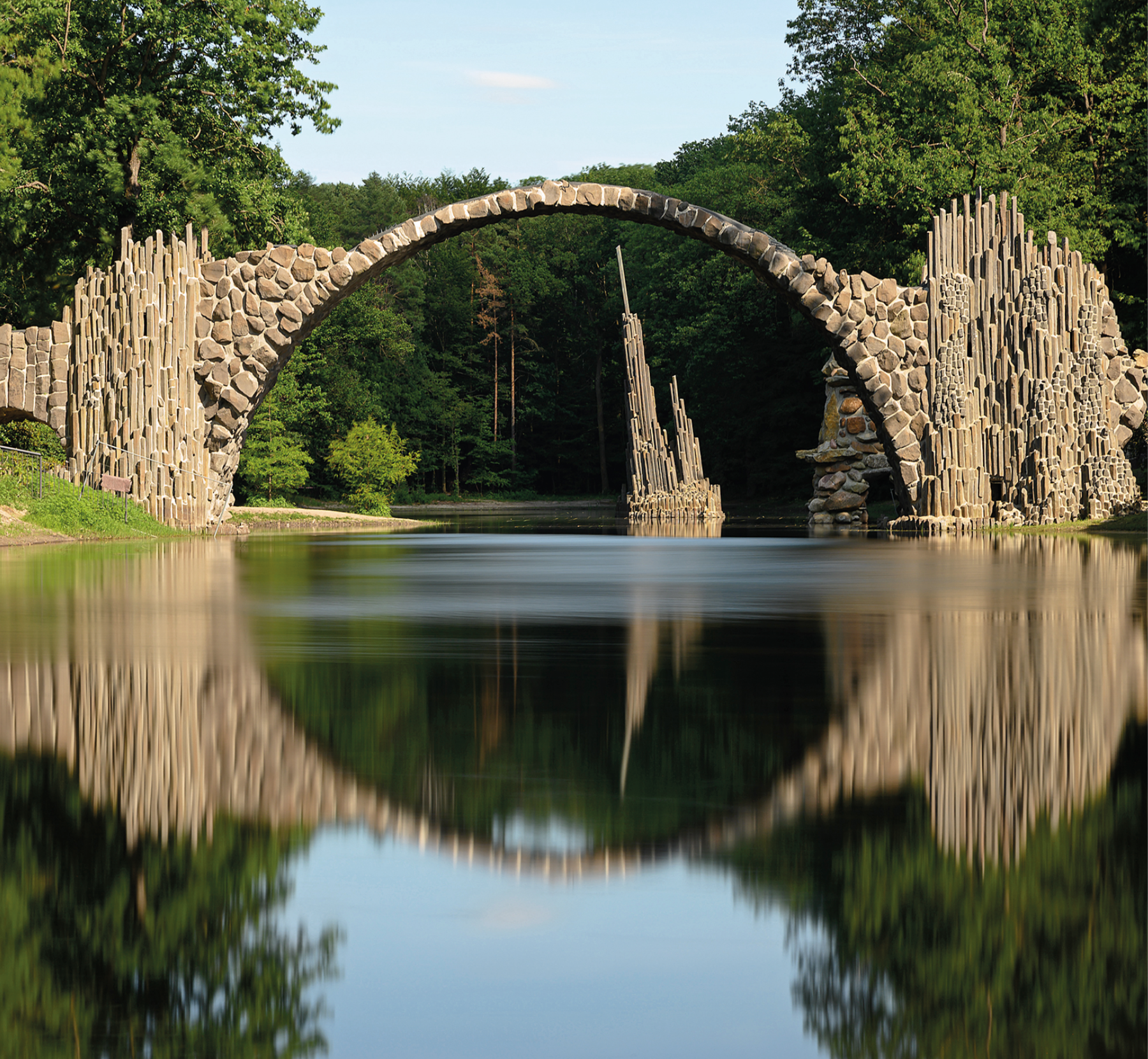 Fotografia.  Ponte do Diabo composto por pedras em formato de arco sobre um rio. No rio, abaixo da ponte, estrutura com hastes de tamanhos diferentes. Ao redor, vegetação e árvores.