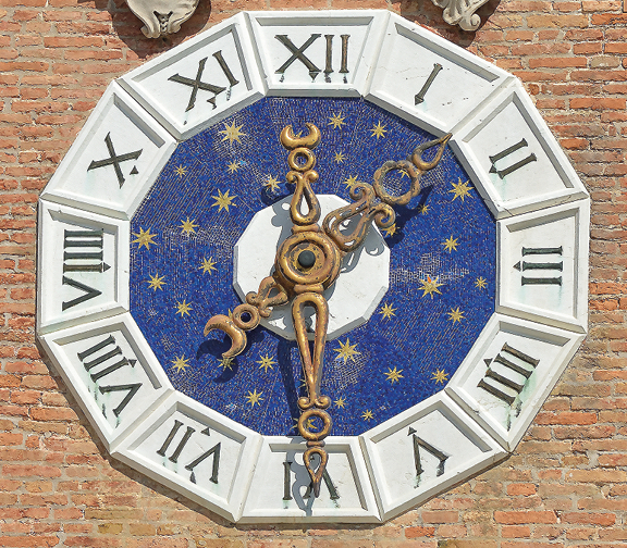Fotografia. Relógio com números romanos e em formato de dodecágono . O fundo é azul e os ponteiros são dourados.
