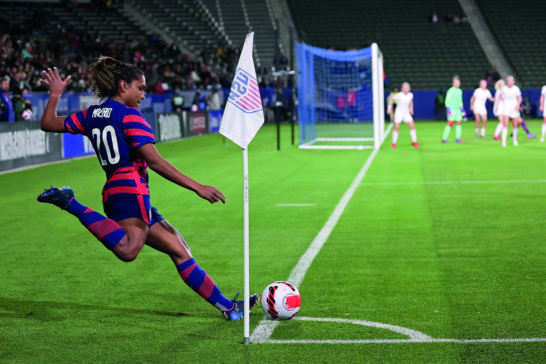 Fotografia. Mulher uniformizada em campo de futebol. Ela cobra escanteio onde há uma bandeira. Ao fundo, pessoas na frente do gol.
