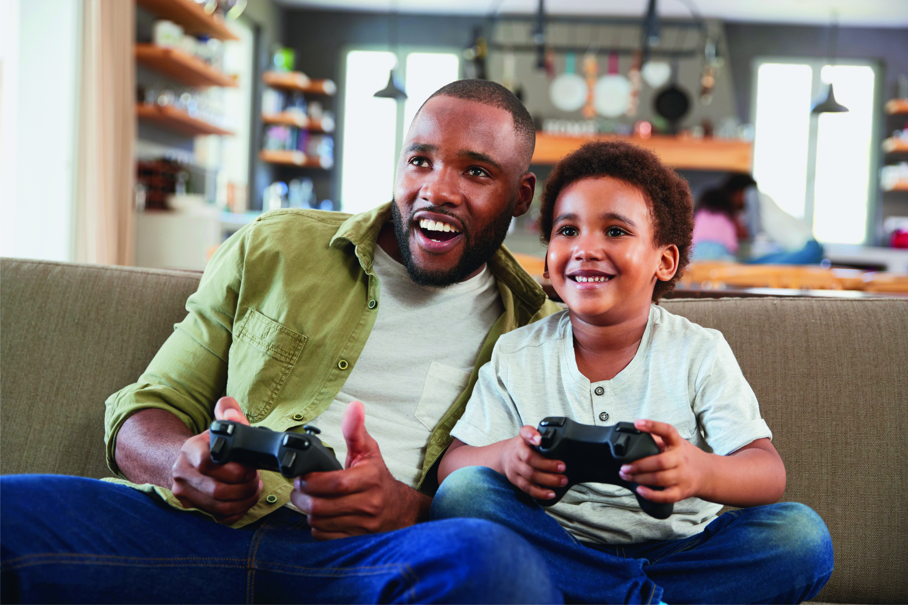 Fotografia. Um homem de cabelo curto, usando uma jaqueta verde, camiseta branca, está sentado em um sofá, segurando um controle de videogame e sorrindo; ao seu lado, um menino de cabelo cacheado e curto, usando camiseta branca, está sentado no sofá, segurando um controle de videogame e sorrindo.