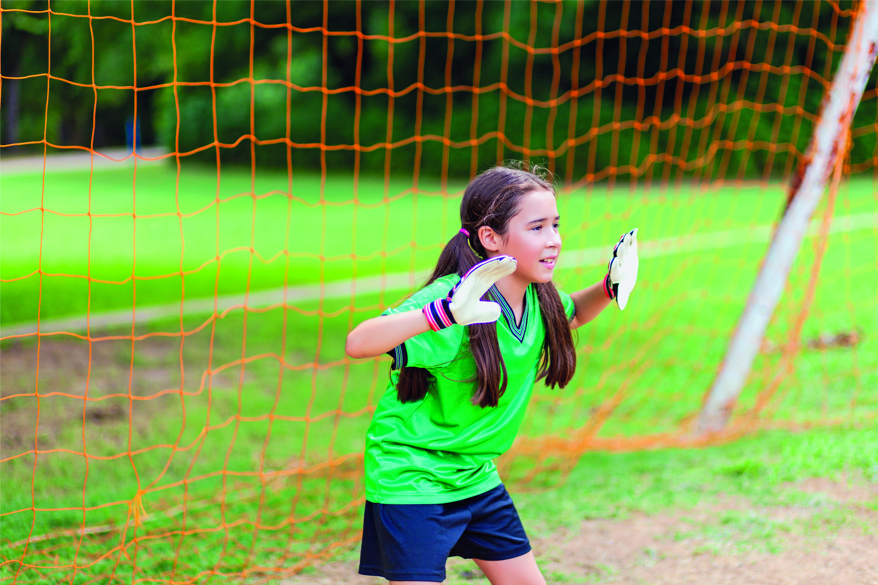 Fotografia. Uma menina de cabelo liso e preso em maria chiquinha, usando um uniforme verde, uma luva branca em cada mão. Ela está na frente de um gol, com as duas mãos espalmadas à frente do corpo.