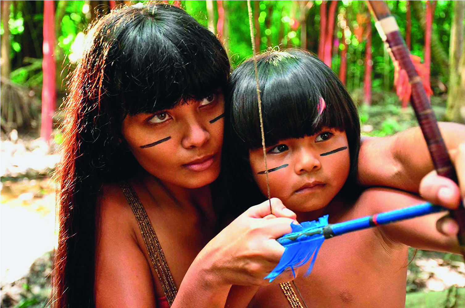 Fotografia. Uma mulher indígena de cabelo preto e liso, com duas linhas pretas pintadas no rosto; ela está com uma criança indígena no colo, também com duas linhas pintadas no rosto. Ambas seguram um arco marrom com uma flecha azul com penas azuis.