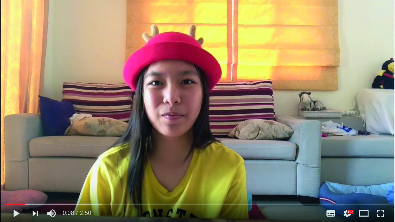 Captura de tela. Uma menina de traços orientais, cabelo liso, usando um chapéu redondo vermelho e camiseta amarela. Ao fundo há um sofá e uma janela.