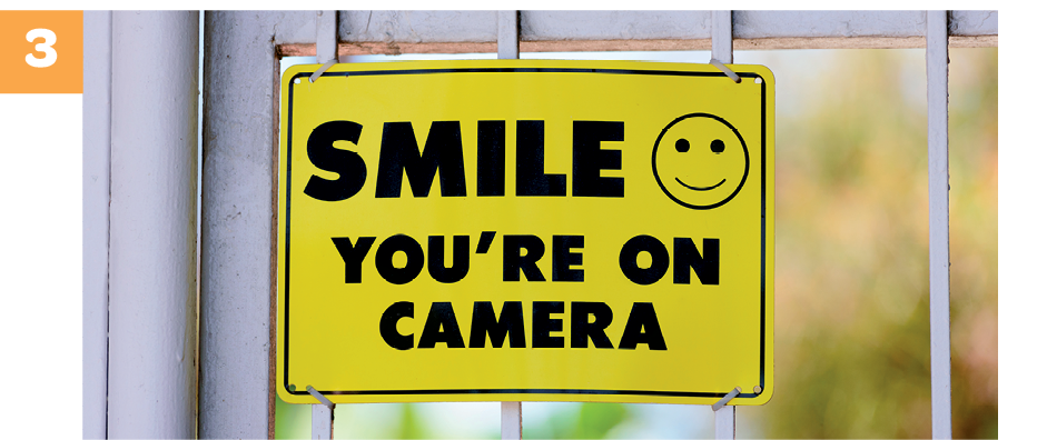 Fotografia. Número 3. Uma placa amarela afixada numa grade. Na placa se lê: SMILE, YOU’RE ON CAMERA. À direita, há um emoji de rosto sorrindo.