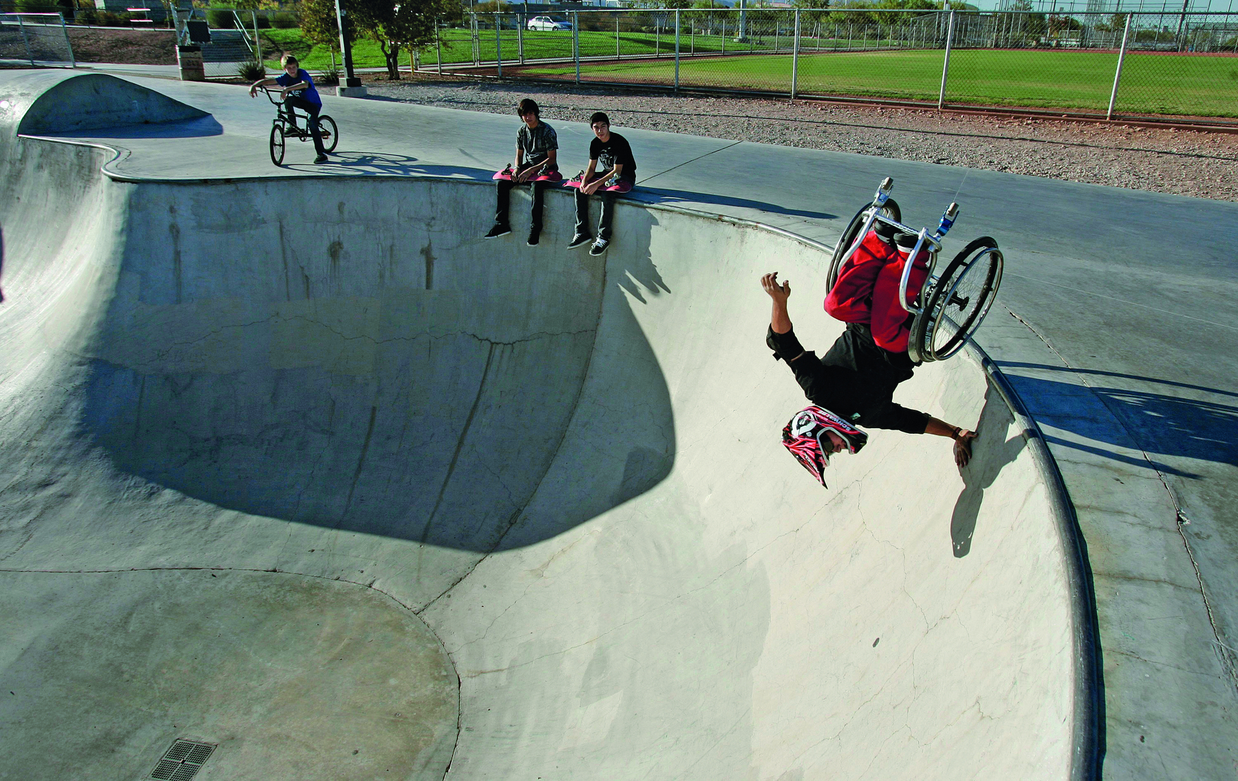 Fotografia. Um homem usando capacete e sentado em uma cadeira de rodas está no topo de uma pista de skate com a mão apoiada na borda e a cadeira com as rodas para o alto. Ao lado, duas pessoas sentadas na borda da pista estão observando.