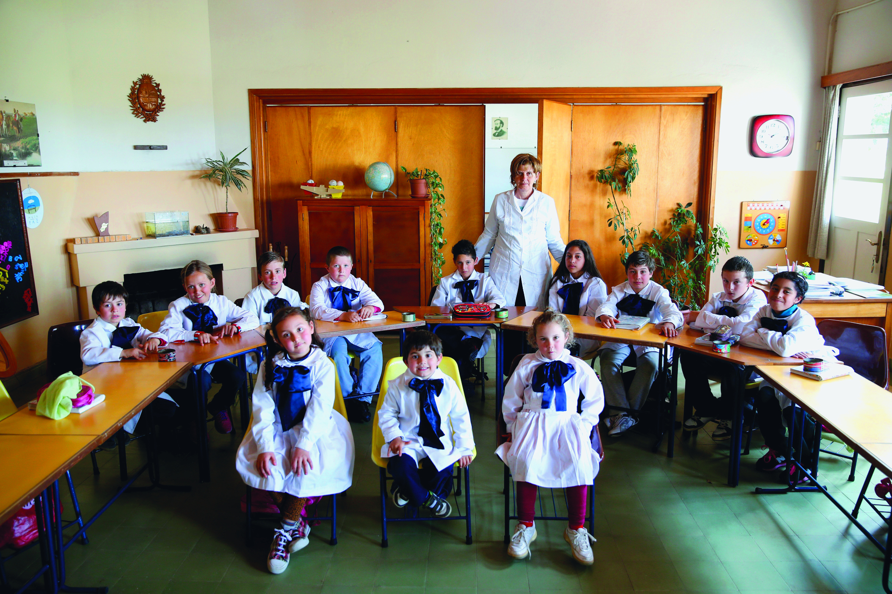 Fotografia. Vista panorâmica de uma sala de aula com mesas de madeira em disposição circular. Crianças brancas usando uniforme branco com um lenço azul estão sentadas; ao fundo, uma professora está em pé. A sala tem armários de madeira ao fundo e algumas plantas.