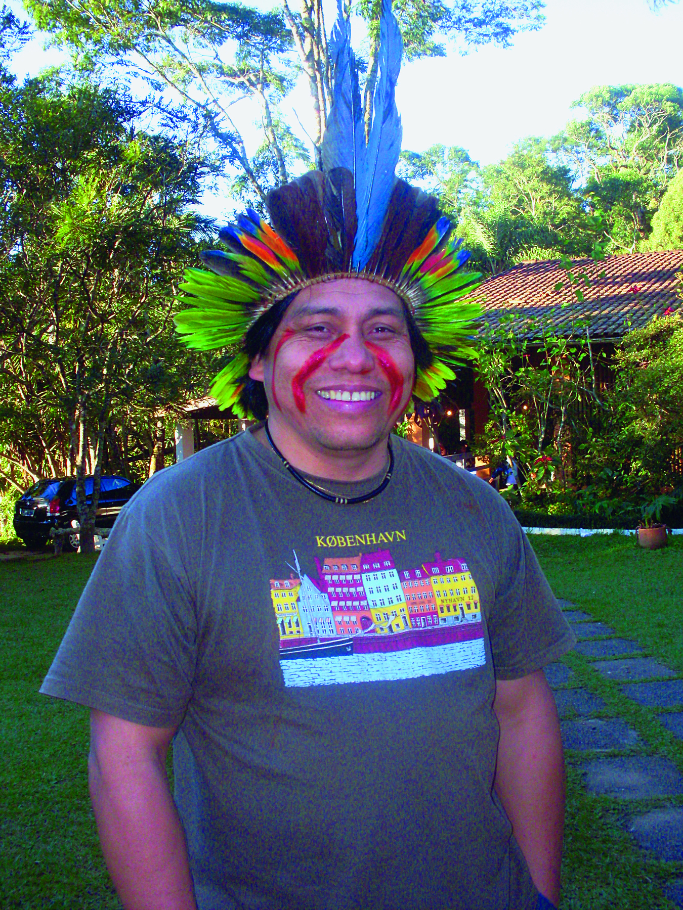 Fotografia. Daniel Munduruku. Um homem indígena, de cabelo preto, usando um cocar feito com penas verdes e pretas, camiseta cinza escuro e com linhas vermelhas pintadas no rosto. Ele está sorrindo.