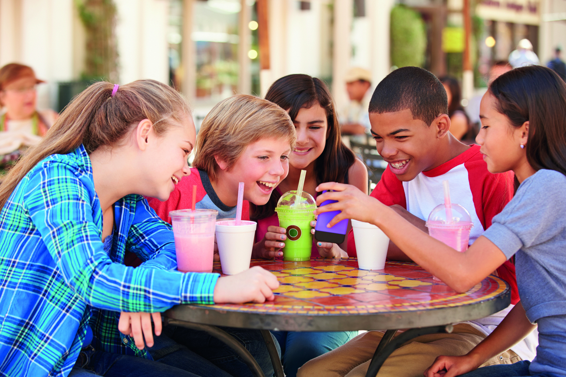 Fotografia. Diversas crianças estão sentadas ao redor de uma mesa redonda. Sobre a mesa há copos com suco. As crianças estão sorrindo e olhando na direção de um celular.