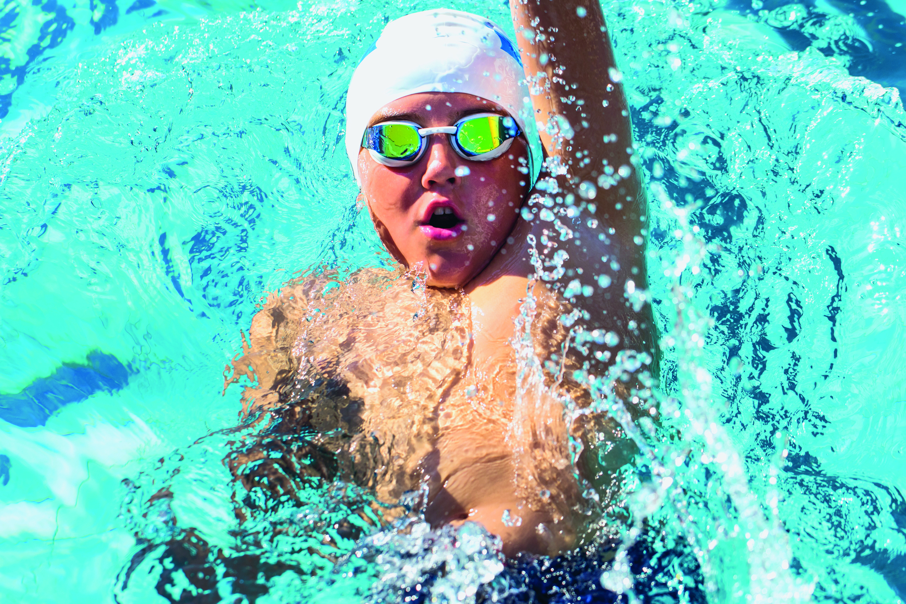 Fotografia. Uma criança com touca e óculos de natação está dentro da piscina, de costas, com o braço esquerdo atrás da cabeça.