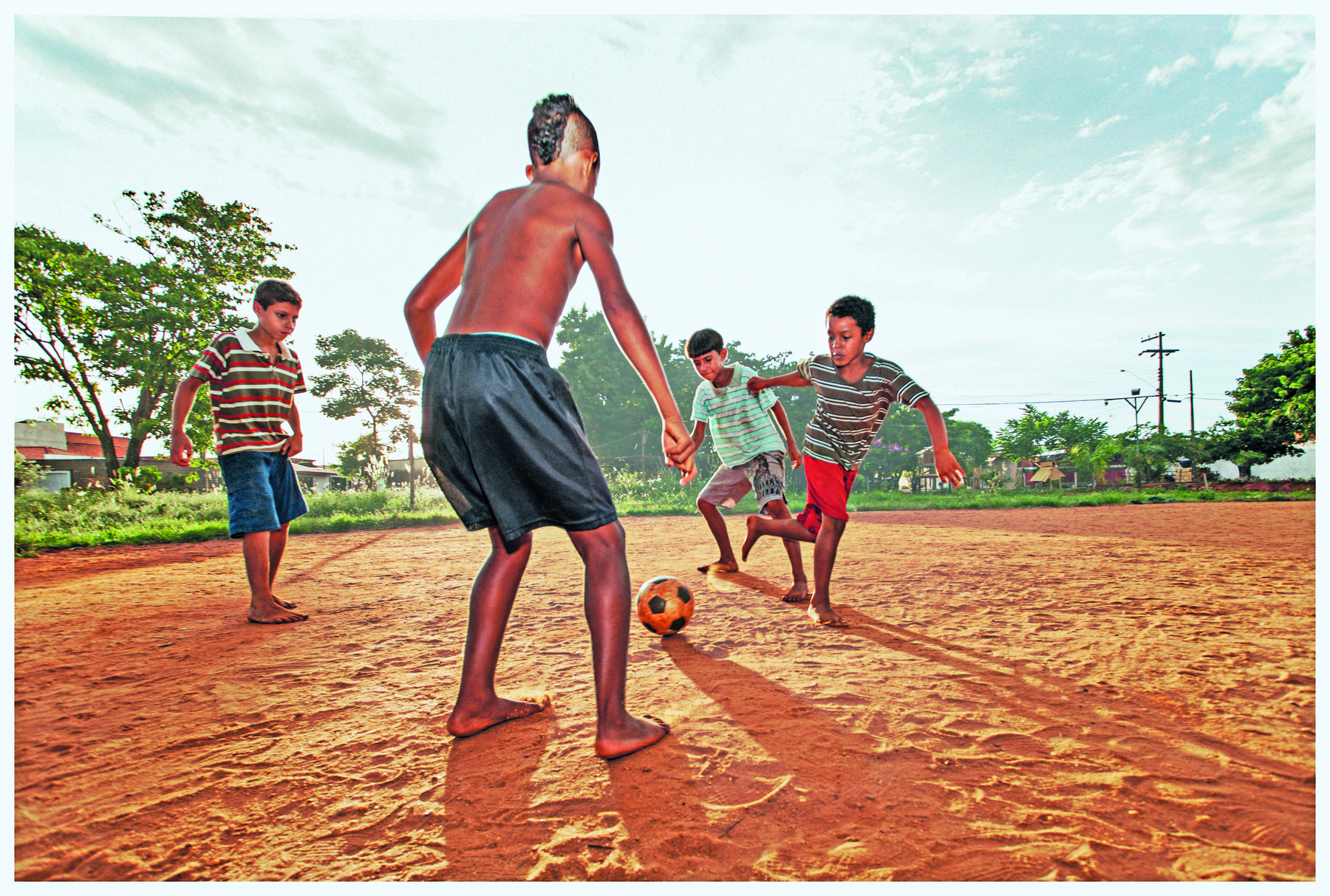 Fotografia. Quatro crianças descalças em um campo de terra. Entre elas há uma bola de futebol.