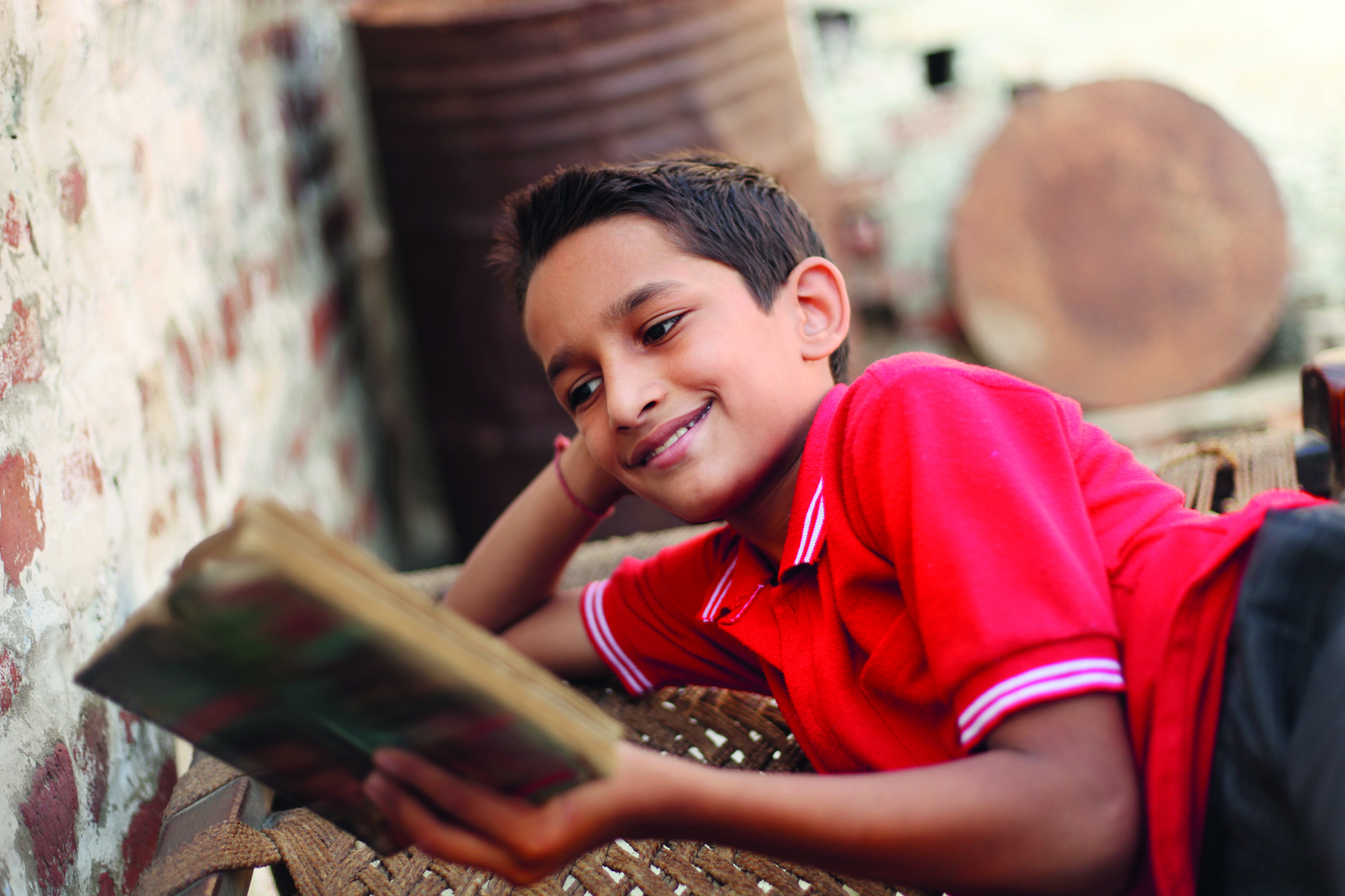 Fotografia. Um menino de cabelo castanho curto usando uma camisa polo vermelha. Ele está deitado de barriga para baixo, com a cabeça apoiada na mão direita, olhando para um livro aberto e sorrindo.