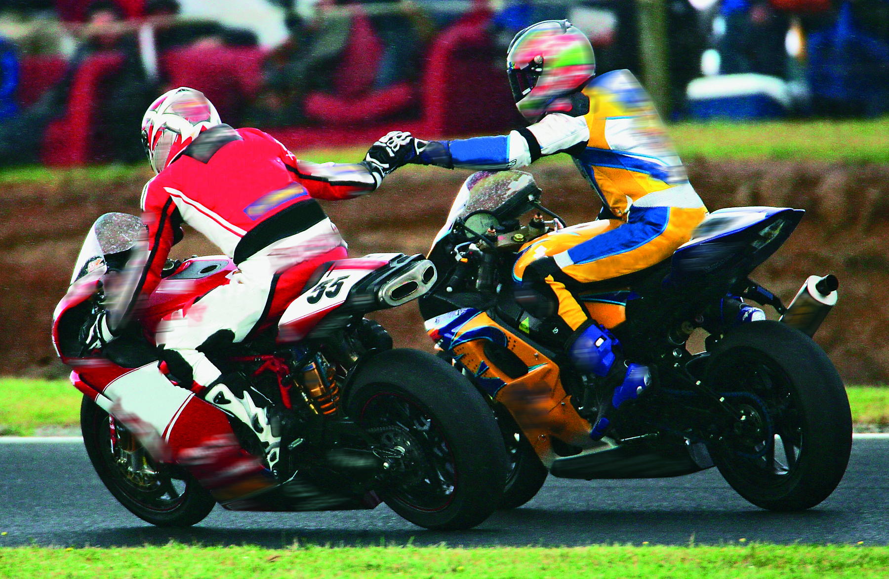 Fotografia. Dois homens usando roupas grossas, capacetes, estão montados em motos grandes. Os dois estão com os braços esticados um na direção do outro, se cumprimentando.