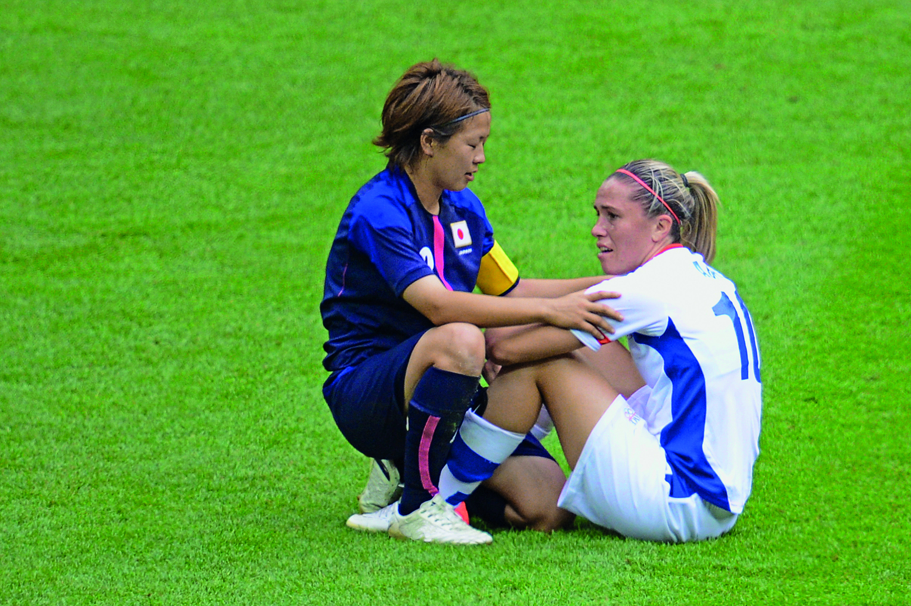 Fotografia. Duas mulheres, uma usando uniforme azul; outra usando uniforme branco, estão sentadas em um gramado de campo de futebol. A mulher de uniforme azul está de frente para a mulher de uniforme branco e com as mãos sobre os seus braços.