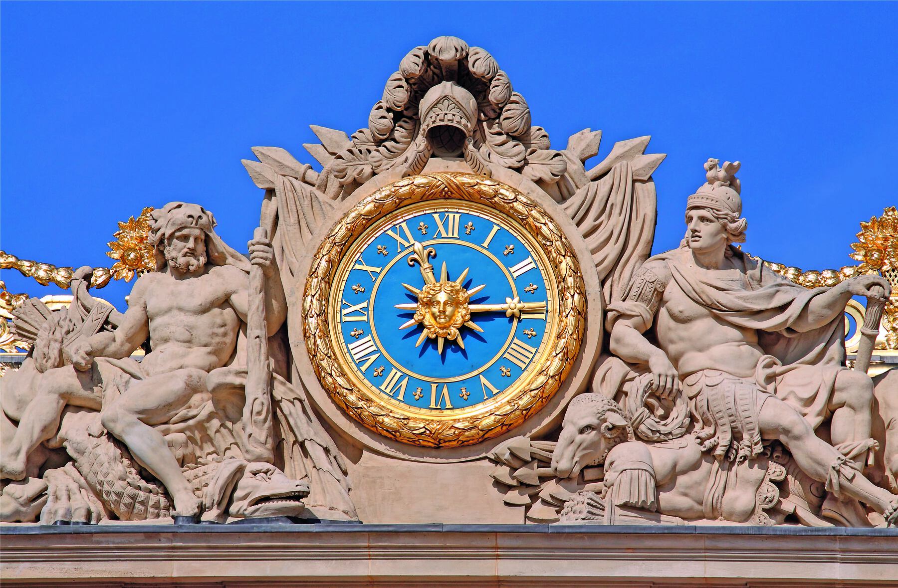 Fotografia. Um relógio de ponteiros dourado com fundo azul. No centro dele há um sol e, nas laterais, há esculturas de pessoas feitas em pedra.