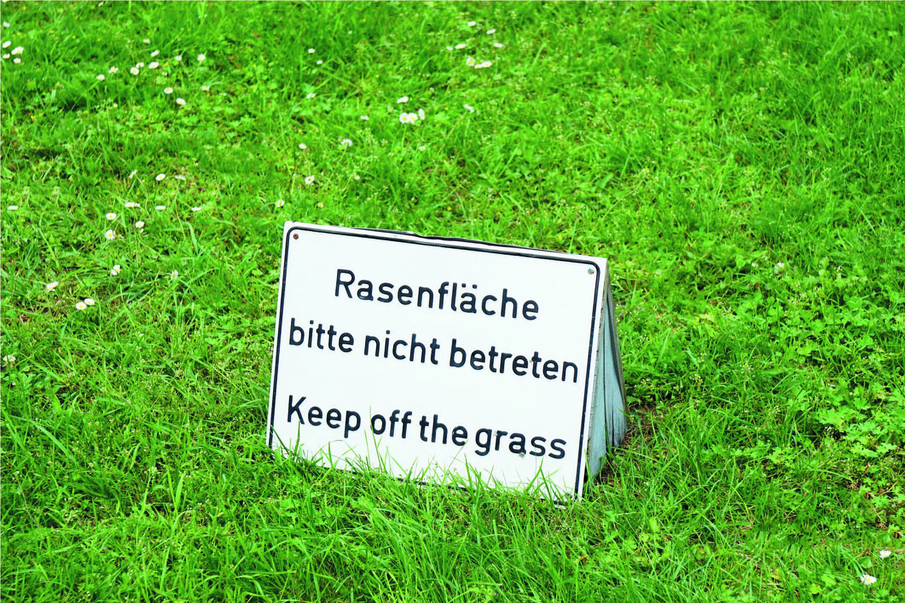 Fotografia. Letra a. Placa branca bilíngue sobre um gramado com os dizeres: RASENFLÄCHE BITTE NICHT BETRETEN. KEEP OFF THE GRASS.