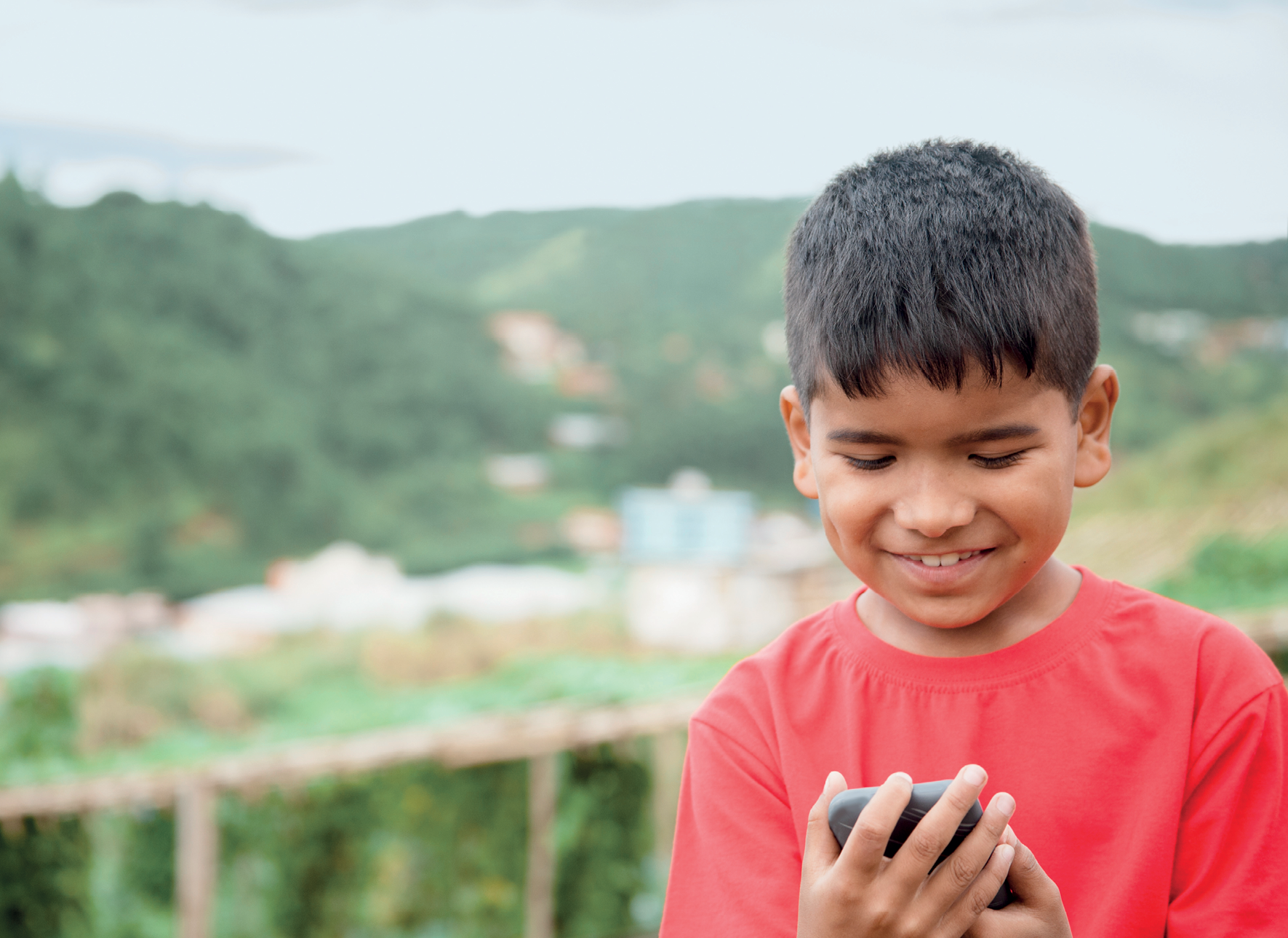 Fotografia. Um menino de cabelo castanho e curto, de sobrancelhas grossas, usando camiseta vermelha, está segurando um celular com as duas mãos, olhando para o celular e sorrindo.