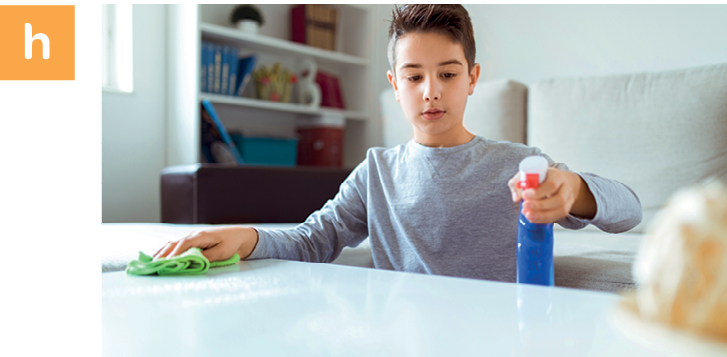 Fotografia. Letra h. Uma criança está segurando um pano sobre uma mesa. Com a outra mão ele está espirrando um produto de limpeza sobre a mesa.