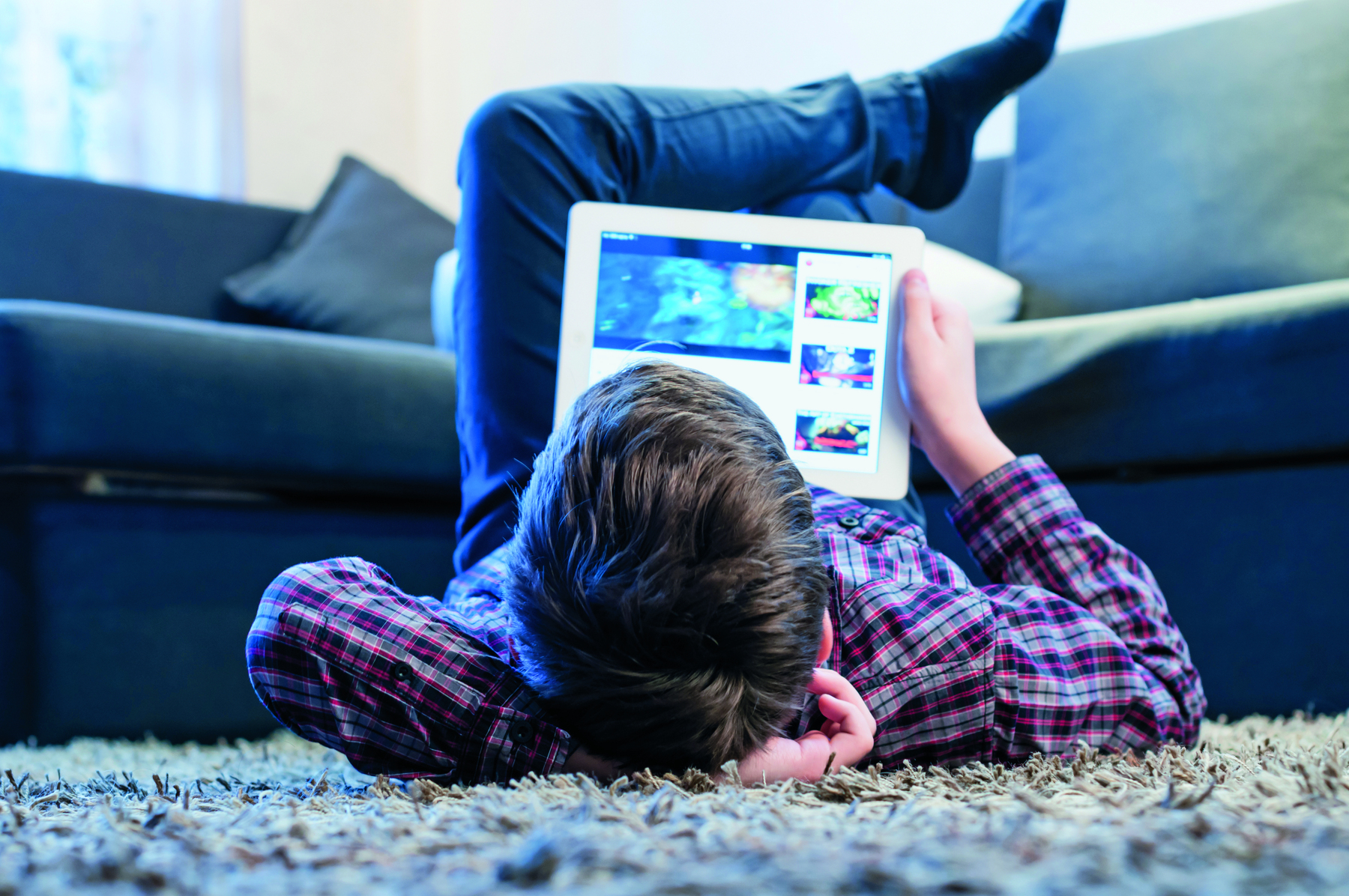 Fotografia. Um menino de cabelo castanho, usando camiseta xadrez está deitado no chão com as pernas cruzadas para cima. Ele está segurando e um tablet branco apoiado na barriga. Na tela do tablet há uma plataforma de streaming de vídeos.