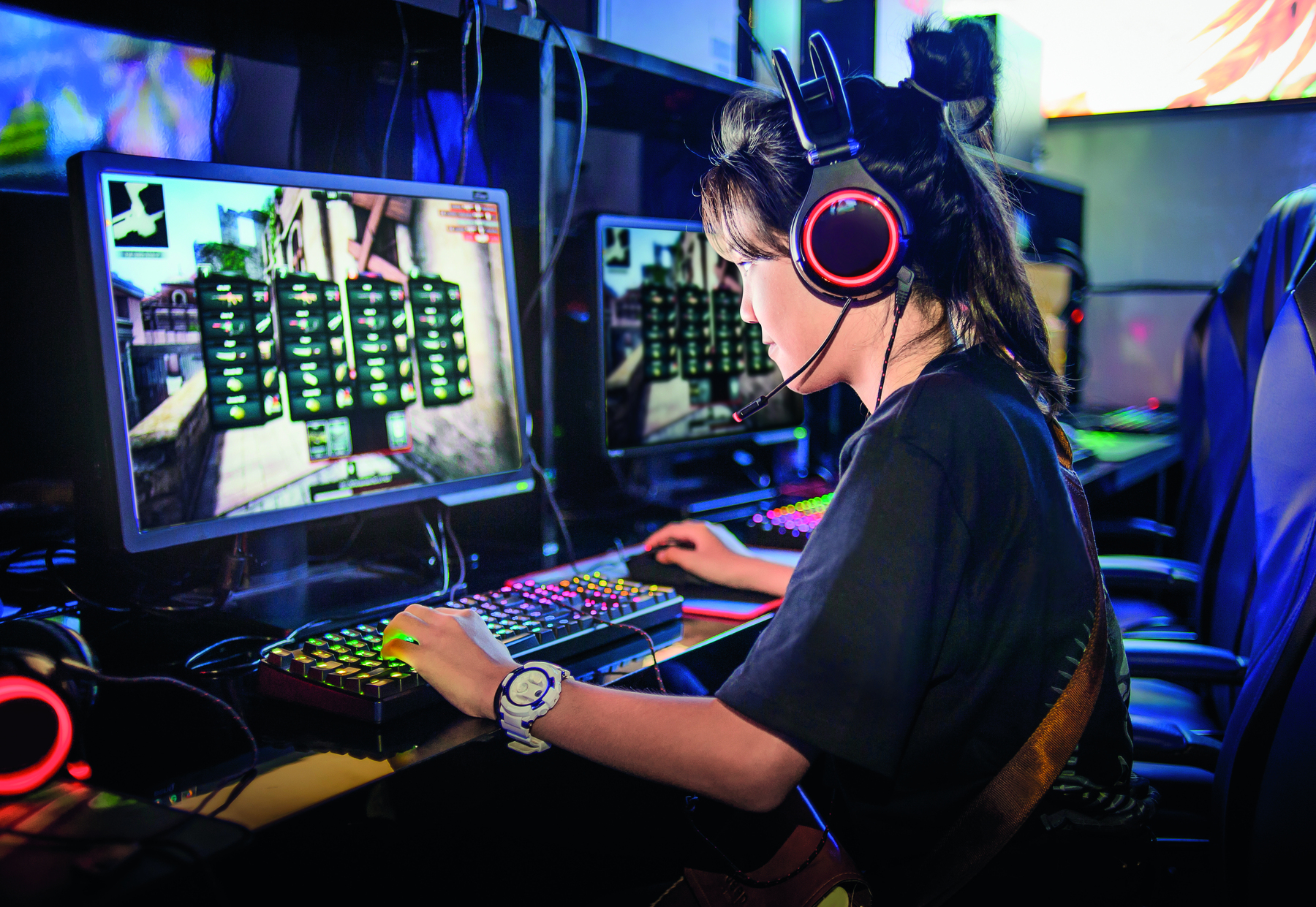 Fotografia. Uma menina de cabelo castanho comprido, usando camiseta preta e fone de ouvido redondo com microfone. Ela está sentada à mesa com as duas mãos na direção de um teclado colorido. Seu rosto está inclinado na direção de um monitor grande. A seu lado há outro teclado e outro monitor grande.