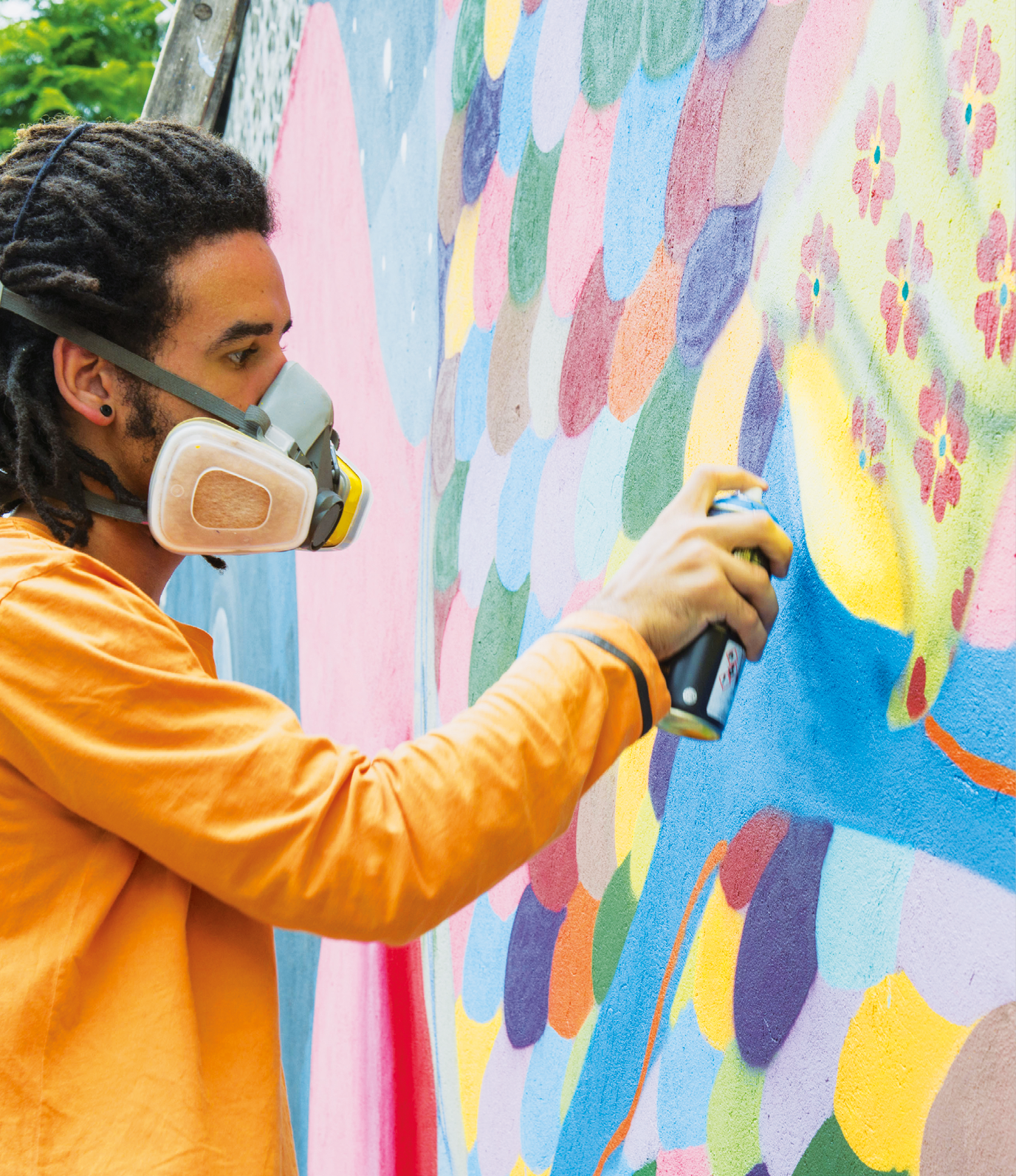 Fotografia. Um homem de cabelo penteado em tranças, usando blusa laranja e uma máscara protetora cinza sobre o nariz e a boca. Ele está segurando uma lata de spray e apontando-a na direção de um muro colorido.