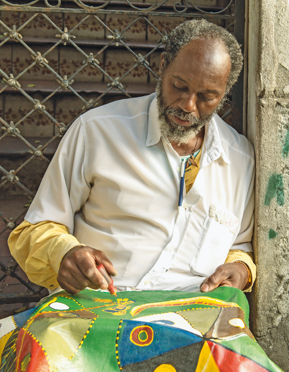 Fotografia. Um homem de cabelo grisalho e cacheado, usando camisa branca. Ele está apoiado em uma parede, segurando uma bisnaga de tinta amarela e despejando um pouco de tinta sobre um pano verde com uma pintura em amarelo, azul, vermelho, branco, verde e preto.