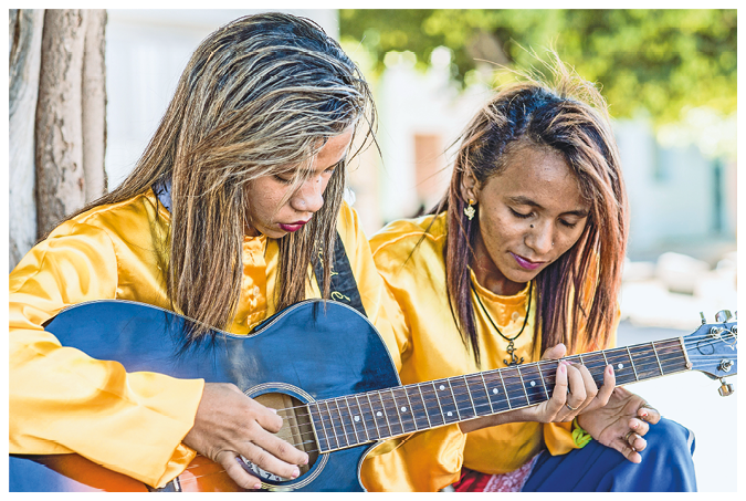 Fotografia. Uma menina de cabelo loiro penteado com tranças usando uma blusa amarela está com a cabeça baixa na direção de um violão preto. Sua mão esquerda está segurando o braço do violão e a mão direita está dedilhando as cordas. A seu lado há uma mulher de cabelo castanho usando uma blusa amarela com a cabeça baixa.