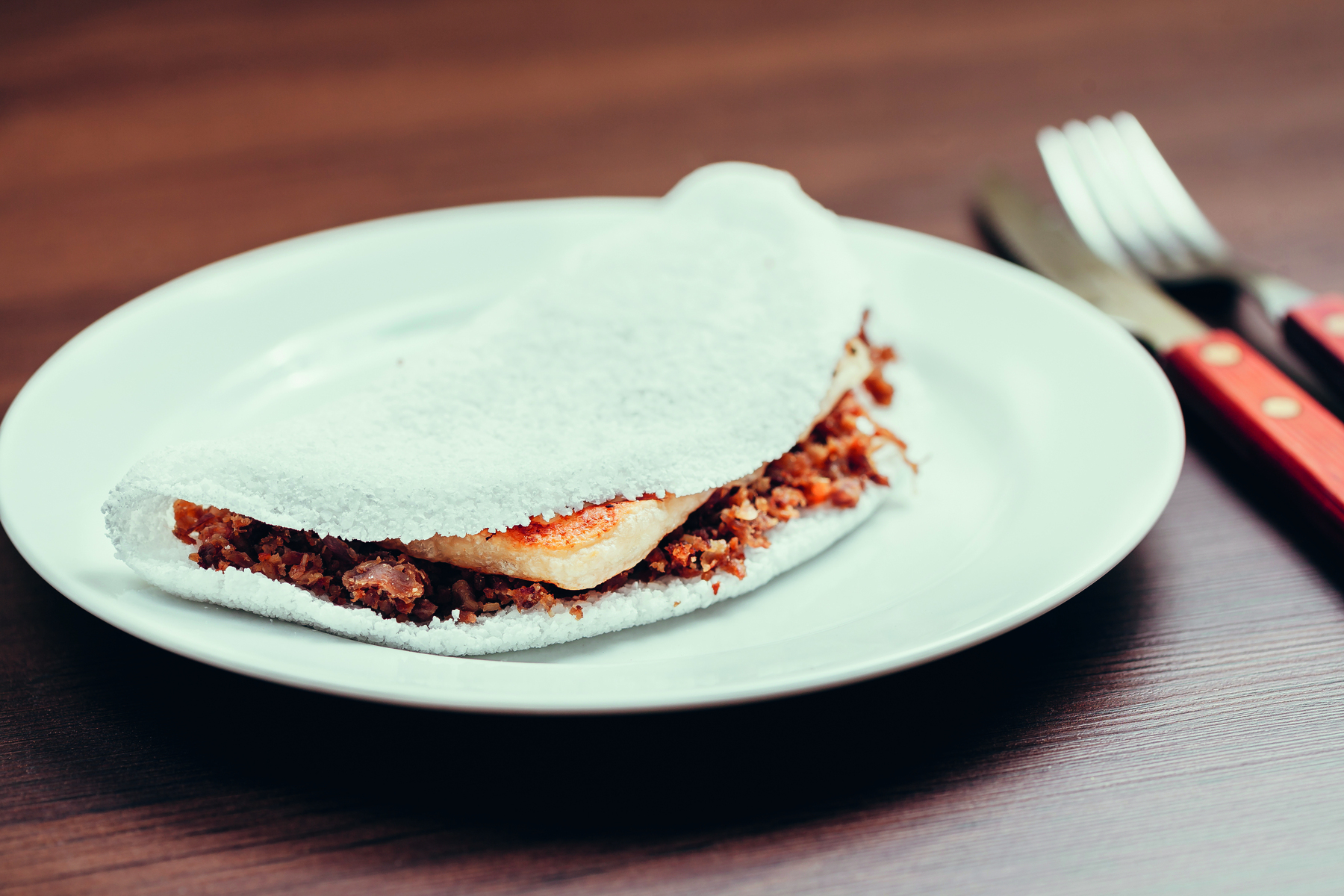 Fotografia. Um prato branco com uma tapioca branca recheada. Ao lado há uma faca e um garfo.