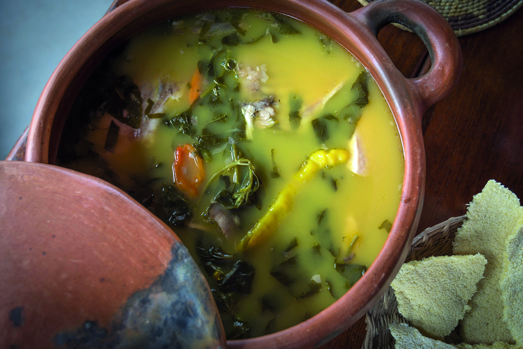 Fotografia. Uma panela marrom com sopa amarela com folhas verdes.