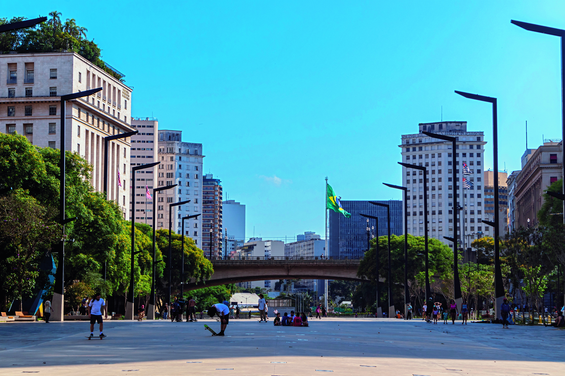 Fotografia. Vista de uma praça larga com postes de luz e árvores nas laterais. No centro há pessoas sentadas e andando de skate. Ao fundo há um viaduto com uma bandeira do Brasil.