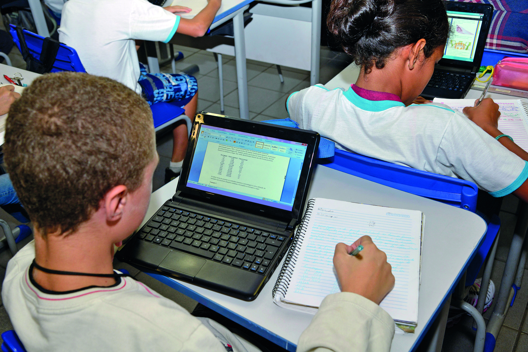 Fotografia. Perfil de um menino loiro usando uniforme branco. Ele está sentado em sua carteira escolar, onde há um laptop preto e um caderno. Ele está segurando uma caneta apontada para o caderno. Ao seu redor há outros alunos sentadas em carteiras.